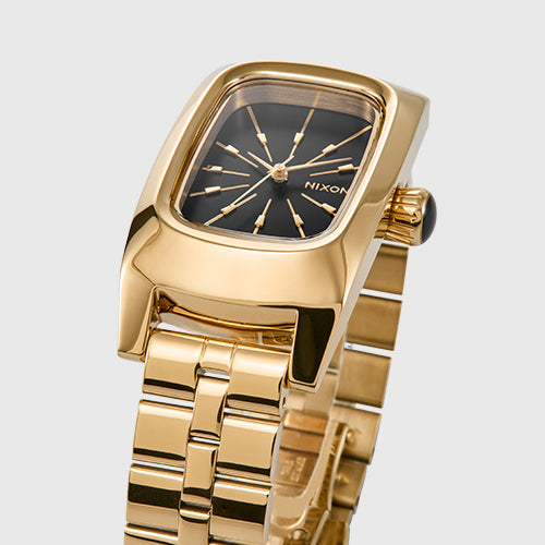 A close-up shot of a gold Nixon watch with a black rectangular face and gold hour markers, featuring a matching gold link bracelet.