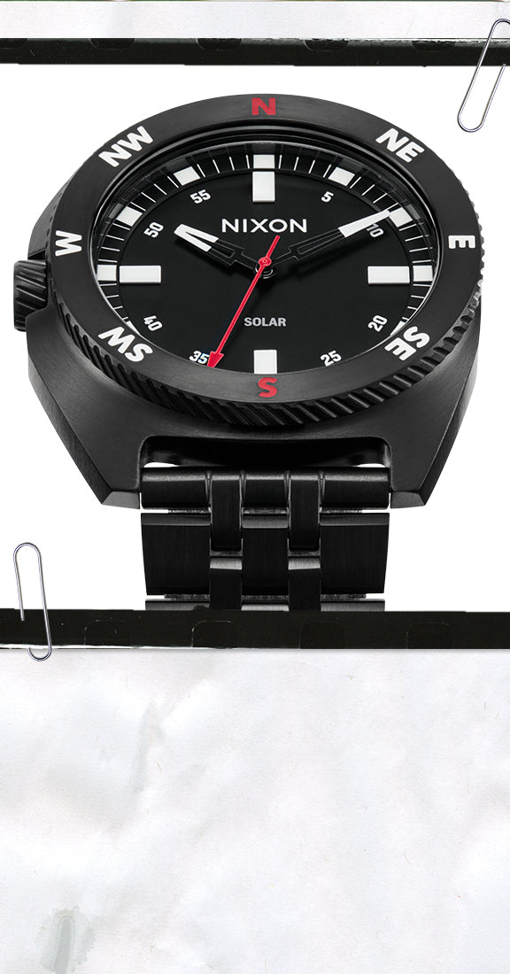 A black Nixon Solar watch with a black dial, black bezel featuring white compass directions, and a black metal link bracelet, shown against a white background with paper clips.