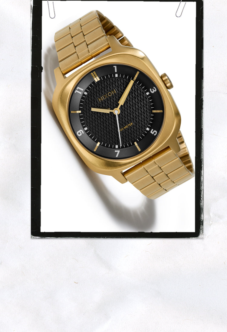 A gold-tone metal wristwatch with a black textured dial and gold-tone bezel details, shown inside a black-bordered white frame on a white background.