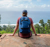 A man sits on a cliff and looks out over the ocean while wearing a blue Nixon backpack.