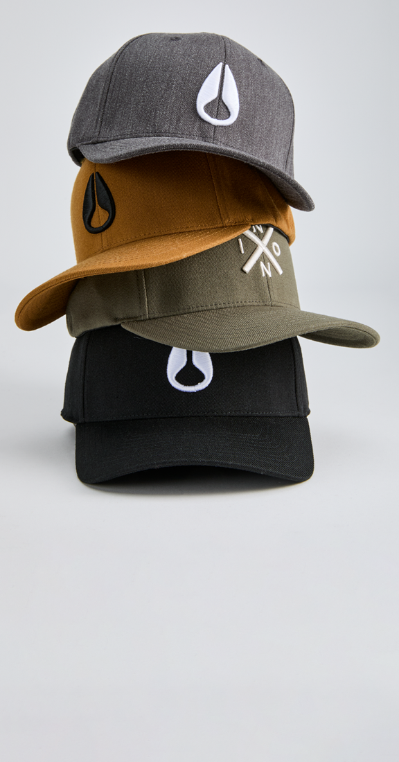 Four snapback caps stacked vertically on a light gray studio background. The caps are heather gray, mustard brown, olive green, and black, each featuring a white Nixon logo on the front or side.